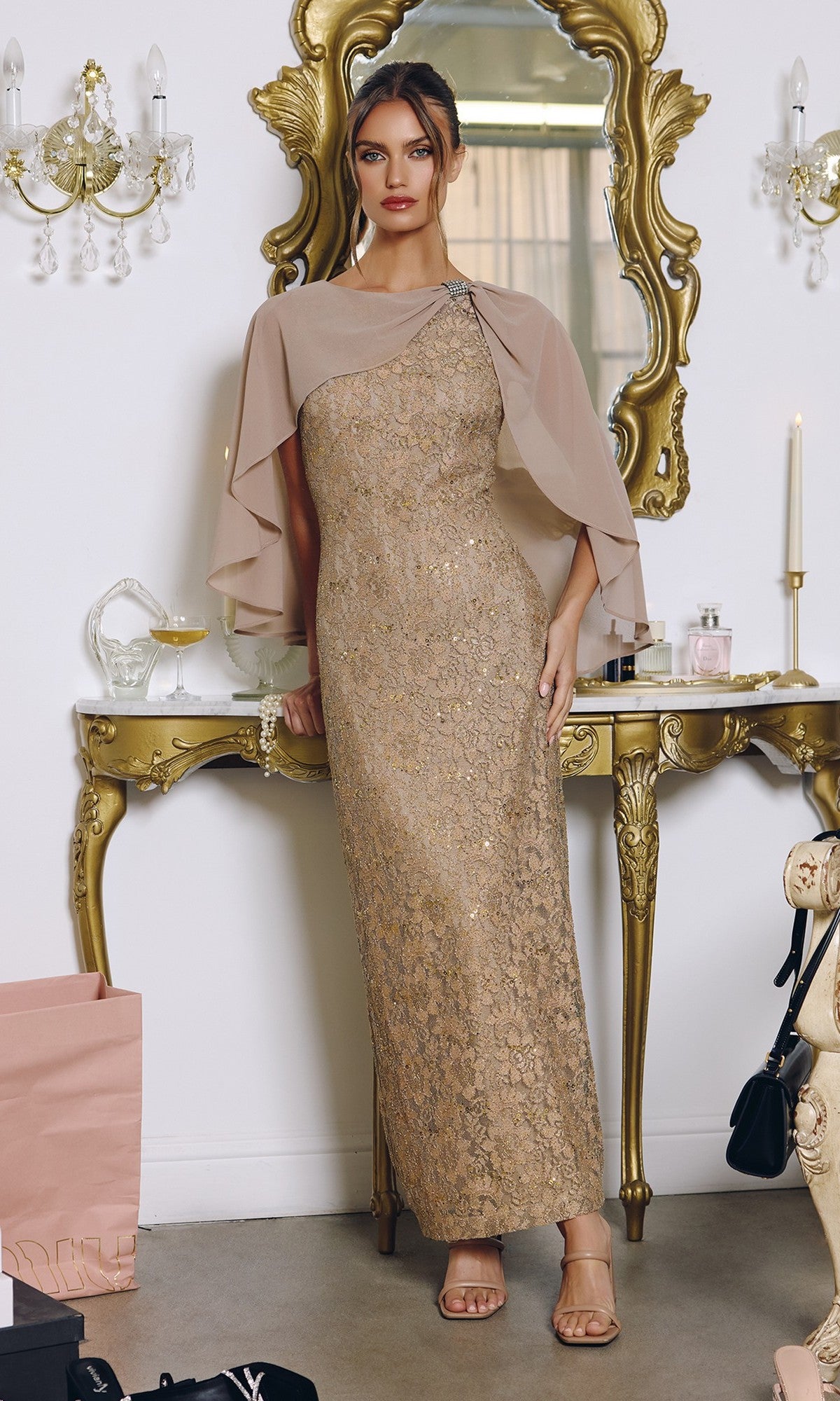 A woman in the Amoris T906 mocha long prom dress, featuring a one-shoulder design and chiffon cape overlay, stands before an ornate gold mirror and table adorned with decorative accents and shopping bags.